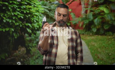 Mann, der einen elektrischen Bohrer in der rechten Hand hält, auf einem Straßenweg, der von grünem Parklaub eingerahmt ist, Bart und Schreinerflanell sichtbar ist; entschlossenes Handwerkskunst Stockfoto
