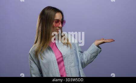 Frau, die eine Geste in einem blauen Hemd mit verspieltem Ausdruck macht, die auf ihre offene Handfläche über einem leuchtend violetten Hintergrund zeigt, mit rosa Sonnenbrillen und Stockfoto