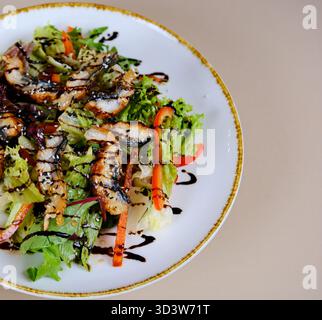 Eine exquisite Präsentation eines Salats mit gegrilltem Aal, frischen Salatblättern und saftigen Kirschtomaten auf einem Teller, leicht bestreut mit Sesamsamen. Ausgestellt auf einem Holztisch. Hohe Qualität Stockfoto