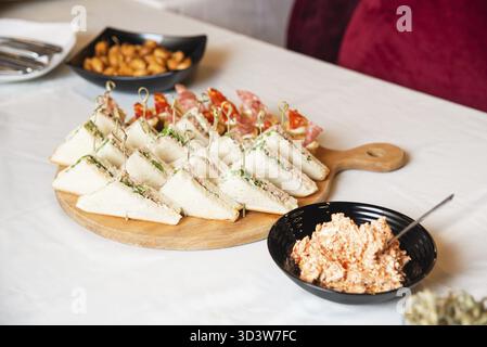 Speisen, verschiedene Dreieck-Sandwiches mit Aufschnitt und Gemüse auf Holzbrett, dazu cremiger Aufstrich und Snacks Stockfoto