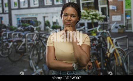 Frau mit Hand am Kinn auf dem Fahrradparkplatz auf der Straße, trägt Armbänder und Jeans und steht mit einem Arm über der Taille; durchdachtes Vertrauen. Stockfoto
