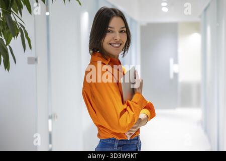 Porträt einer lächelnden Geschäftsfrau mit dem Laptop in den Händen, der im Korridor des Büros steht Stockfoto