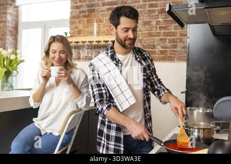 Glückliche attraktive junge Frau, die ihren Mann unterstützt, während er ein Abendessen für die Familie kocht, eine Frau, die Kaffee trinkt Stockfoto
