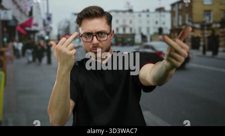 Junger bärtiger Mann auf einer Stadtstraße, der eine unhöfliche Handbewegung mit beiden Mittelfingern macht, während er Brille und ein schwarzes Hemd in einem urbanen Outd trägt Stockfoto