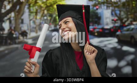 Hispanische Frau in Abschlusskleidung lächelt fröhlich auf einer Stadtstraße mit einem Diplom in der Hand vor einem Hintergrund der städtischen Aktivität und des hellen Tageslichts. Stockfoto