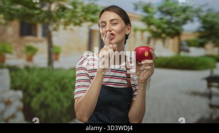 Frau, die roten Apfel mit Handschuhen vor dem Gebäude im Freien hält, in Richtung Kamera lächelt; frische gesunde Snackfreude. Stockfoto