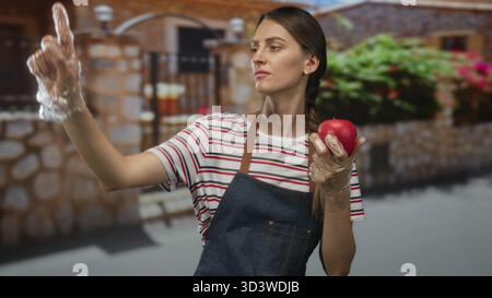 Frau, die einen roten Apfel mit Handschuhen am Eingang des Gebäudes in Schürze und gestreiftem Hemd hält; Ekel. Stockfoto