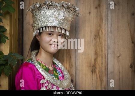 Feng Huang, China - August 2019 : schöne Chinesin in traditioneller Volkstracht gekleidet und posiert für ein Bild vor hölzernem Hintergrund von an Stockfoto