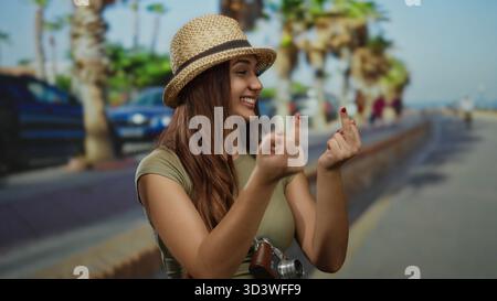 Frau lächelt draußen und macht Gesten in der sonnigen Straße mit Palmen, die Hut und lässige Kleidung tragen und während des Urlaubs verspielte Touristenmanieren zeigen. Stockfoto
