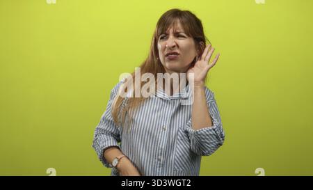 Junge Frau, die Hand an Ohr hält und knisternd, gestreiftes Hemd, limgrün Studio; Verwirrung beim Zuhören. Stockfoto