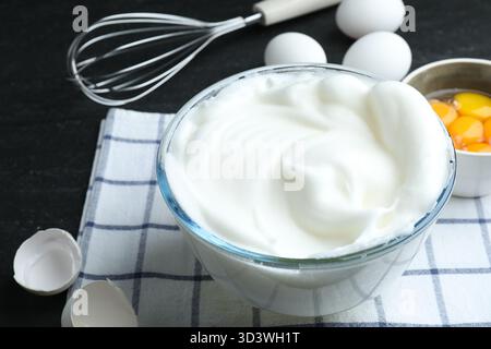Eiweiß in Schüssel, Schneebesen und Zutaten auf schwarzem Tisch, Nahaufnahme Stockfoto