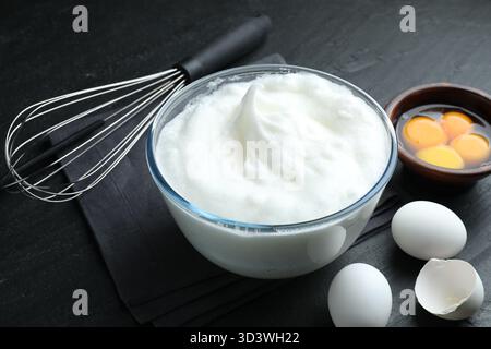 Eiweiß in Schüssel, Schneebesen und Zutaten auf schwarzem Tisch, Nahaufnahme Stockfoto