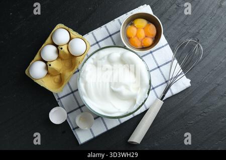 Eiweiß in Schüssel, Schneebesen und Zutaten auf schwarzem Tisch, flach gelegt Stockfoto