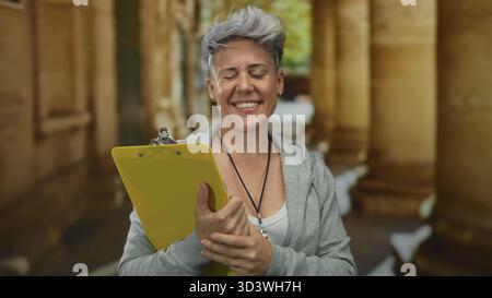 Frau mit kurzen grauen Haaren trägt einen grauen Hoodie und hält das gelbe Klemmbrett mit beiden Händen auf der Straße; Selbstvertrauen. Stockfoto