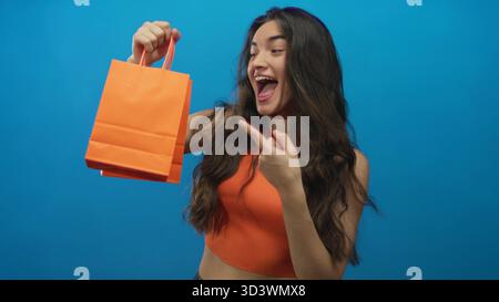 Die junge hispanische Frau lächelt und zeigt den Finger auf die orangefarbene Einkaufstasche im Studio; Aufregung. Stockfoto