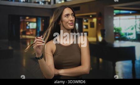 Die Frau wirbelt mit den Fingern in der Hotellobby, wobei ein Arm über die Brust gefaltet ist; selbstbewusste, ungezwungene Haltung. Stockfoto