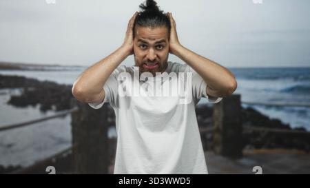 Der Mann bedeckt die Ohren mit Händen am Straßenpier am Meer, hält den Kopf und grimmt; überwältigte Frustration. Stockfoto