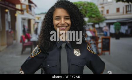 Junge hispanische Polizistin in Uniform lächelt selbstbewusst auf einer belebten Straße draußen mit Leuten und Cafés im Hintergrund. Stockfoto