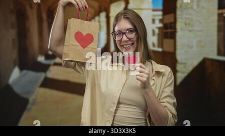 Frau in Brille hält Paperbag mit rotem Herz und Herzausschnitt im Gebäudeflur, während sie lächelt; Liebesfreude. Stockfoto