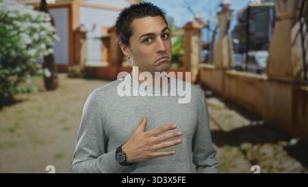 Der junge Mann steht draußen in einer Parklandschaft und drückt seine Kontemplation mit Hand auf Brust aus, vor einem Hintergrund von Bäumen und einem sonnigen Tag. Stockfoto