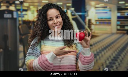Die lächelnde Frau hält das Herz, während sie im Fitnessstudio mit einem farbenfrohen Pullover die Daumen nach oben drückt und im Fitnesscenter positiver wirkt. Stockfoto