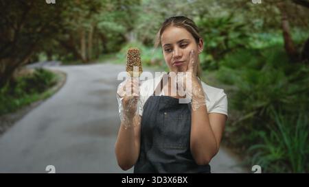 Frau mit Eis und Schürze mit Einweghandschuhen, Finger an Wange auf einer Waldstraße; Versuchungszweifel. Stockfoto