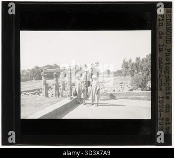 Ein Glasdiapositiv aus der Zeit um 1924-1925, das den Standort des Tempels von Montu nach Abschluss der Ausgrabung zeigt. Im Vordergrund stehen drei Figuren, im Hintergrund die archäologische Stätte. Stockfoto