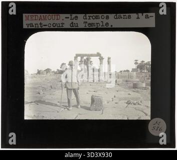Ein Glasdiapositiv aus der Zeit um 1924-1932 zeigt einen Archäologen, der auf den Dromos des Tempels von Montu posiert, der vor dem Innenhof des Tempels steht und einen Moment der Erkundung und Entdeckung festnimmt. Stockfoto