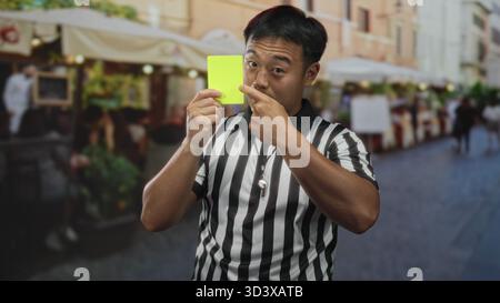 Mann Schiedsrichter in gestreiftem Hemd hält gelbe Karte und zeigt mit dem Finger auf die Straße, während die Restaurantterrasse verschwommen ist; Ernsthaftigkeit. Stockfoto