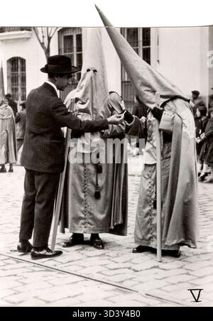 Im Jahr 1929 bietet ein Mann Mitgliedern einer Bruderschaft eine Zigarette an, die während einer Osterprozession in Sevilla, Spanien, spitze Kapuzenmasken tragen. Die Zigarette kann wegen des formalen Charakters der Prozession nicht geraucht werden. 1929, Sevilla, Spanien. Stockfoto