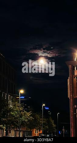 Nächtliche Stadtlandschaft mit leuchtendem Vollmond, umgeben von Wolken, sanftes Licht auf einer ruhigen Straße, gesäumt von Bäumen und Laternenpfosten, Stockfoto