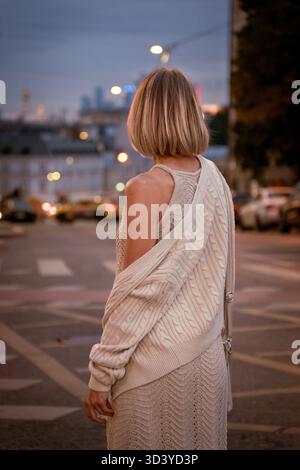 Eine Frau in einem beigefarbenen Strickkleid steht in der Abenddämmerung auf einer Stadtstraße, mit Blick von der Kamera unter warmen Straßenlaternen Stockfoto