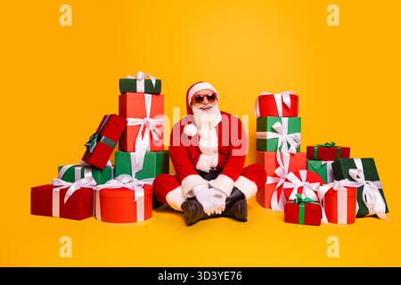 Der Weihnachtsmann sitzt zwischen farbenfrohen Geschenkboxen und trägt einen roten Anzug und eine Sonnenbrille, die für Weihnachtsfotos posiert Stockfoto