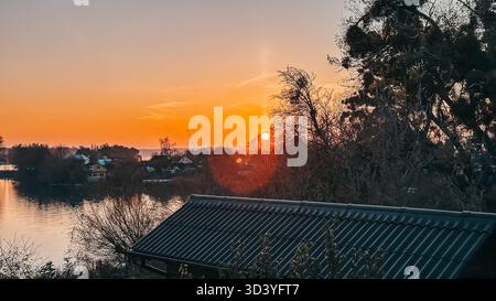 Malerische Aussicht auf einen Sonnenuntergang, der den Himmel in warmen Tönen über einem friedlichen See beleuchtet, umgeben von Silhouetten von Bäumen und Dächern, die eine ruhige und pi schaffen Stockfoto