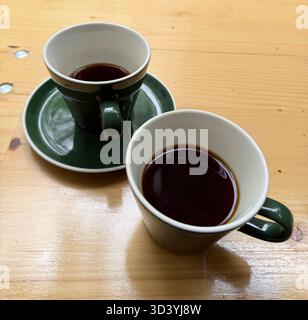 Zwei grüne Keramikbecher mit schwarzem Kaffee auf einem lackierten Holztisch, natürliches Licht, minimales Stillleben. Stockfoto