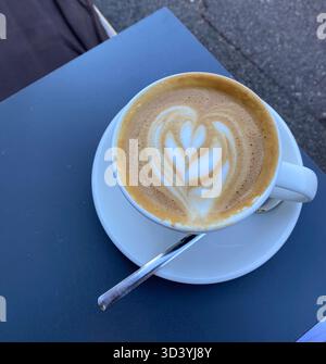 Tasse Cappuccino mit Herz Latte Art in einer weißen Tasse und Untertasse mit Löffel auf einem Café-Tisch im Freien, Blick von oben. Stockfoto