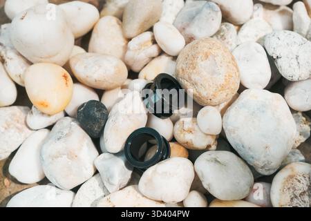Schwarze Kameraobjektivkappen werden auf einem Bett aus glatten Kieselsteinen platziert, die eine natürliche Umgebung im Freien darstellen, die durch Sonnenlicht beleuchtet wird und Texturen hervorhebt Stockfoto