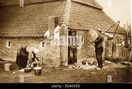 Auf der Insel Terschelling steht 1923 ein Bauer in einer Tür und spricht mit einem Nachbarn, während eine Frau draußen Eimer schrubbt. Auf dem Boden sind Clogs zu sehen, und die Frau trägt eine traditionelle weiße Mütze. Stockfoto