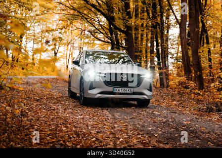 Beige Mazda CX-60 im Herbstwald. Vorderansicht des modernen japanischen SUV auf unbefestigter Straße im Laubwald. Stockfoto