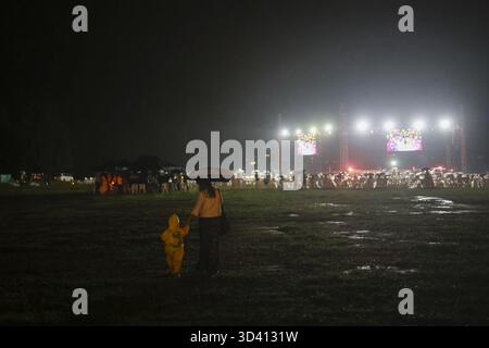 Herzerwärmende Szene der Familie beim Nachtkonzert bei schlechtem Wetter. Eltern und Kind in Regenmantel gehen durch matschiges Feld unter einem Schirm in Richtung helle Straße Stockfoto