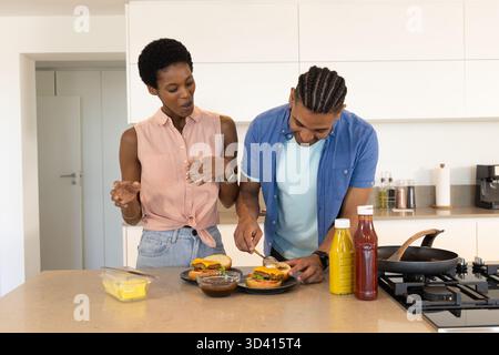 Zubereitung diverser Paare, die Burger auf der Kücheninsel zusammenstellen, mit Soße-Schüssel und Flaschen Stockfoto