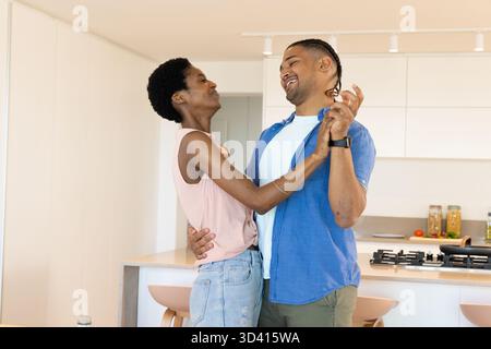 Tanzende, vielfältige Paare halten sich in moderner Küche in der Nähe einer Holzinsel und eines Gaskochfeldes Stockfoto