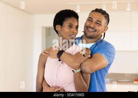 Umarmt verschiedene Paare, die in einer modernen Küche stehen und Smartwatches und weiße Schränke zeigen Stockfoto