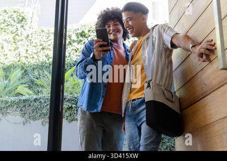Verschiedene männliche Freunde, die durch eine Glastür am Hauseingang schauen, mit Smartphone Stockfoto