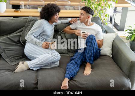 Entspannende männliche Partner, die Kaffeetassen auf grauem Sofa im Wohnzimmer halten, mit Topfpflanzen Stockfoto