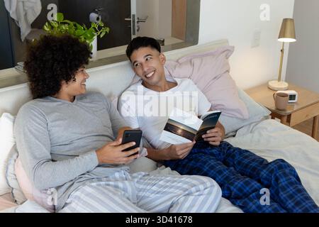 Unterhalten Sie sich mit verschiedenen männlichen Freunden auf dem Bett im modernen Schlafzimmer, mit Smartphone und offenem Buch Stockfoto