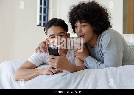 Lachende männliche Freunde, die im Schlafzimmer auf dem Bett liegen und auf das Smartphone schauen, mit weißen Laken Stockfoto