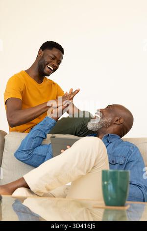 High-Five afroamerikanischer Vater und Sohn sitzen auf Sofa im Wohnzimmer, mit Tablet und Becher Stockfoto