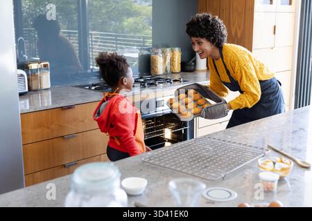 Sie backen verschiedene Mutter und Tochter in Schürzen in der modernen Küche und halten ein Tablett mit Muffins Stockfoto