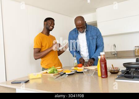 Kochen afroamerikanischer Vater und Sohn, die Burger auf der Kücheninsel machen, mit Smartphone Stockfoto